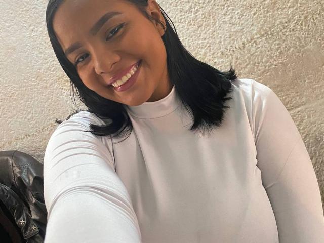 SilverLuna with black hair, smiling, wearing a white long-sleeve turtleneck, showing her teeth, holding a phone for a selfie, and a black leather bag is partly visible.
