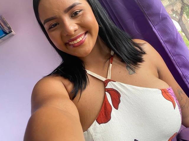 SilverLuna with medium brown skin, black shoulder-length hair, smiling, wearing a white halter top with red floral patterns, showing her big tits and tattoo on her right arm.