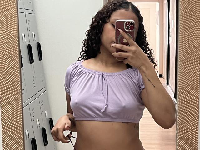 SofiaPassionne with curly dark hair, wearing a lilac crop top, showing her small tits and a tattoo on her right arm, taking a mirror selfie in a locker room.