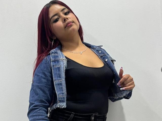 RominaRose with red hair, wearing a denim jacket over a black tank top, showing her tits, black pants, and a necklace, looking down with a slight smirk.
