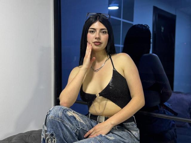 Isabella Rossi mit schwarzen Haaren, die ein schwarzes Crop-Top mit sichtbarer Brust und blaue Jeans trägt, sitzt mit der Hand am Kinn, im Hintergrund ist ihr Spiegelbild zu sehen, und sie trägt eine Brille auf dem Kopf..