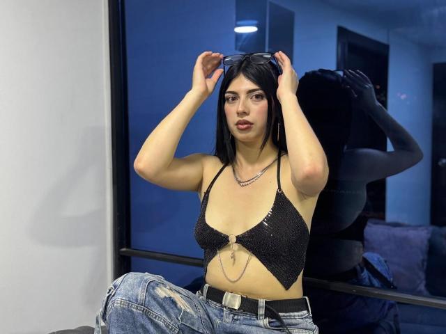 Isabella Rossi, com cabelo preto, vestindo uma cropped preta brilhante, segurando óculos de sol acima da cabeça, mostrando os seios, usando calças jeans rasgadas, em frente a um espelho..