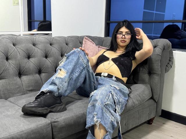 Isabella Rossi, avec ses cheveux noirs, ses lunettes et son ventre légèrement bronzé, porte un crop top noir et un jean déchiré, et est assise sur un canapé gris capitonné, avec des bottes noires..
