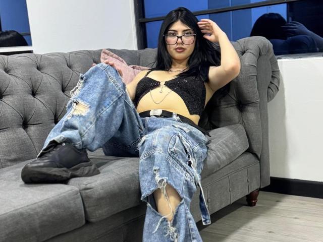 Isabella Rossi, com cabelo preto e óculos, está sentada em um sofá cinza com detalhes em tufos, vestindo uma cropped preta com glitter, jeans azuis rasgados e botas pretas, com a mão no cabelo..