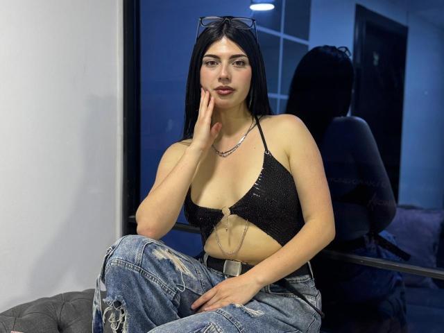 IsabellaaRossi with black hair, wearing a glittery black crop top, showing her tits, and blue ripped jeans, sitting, hand on cheek, mirror reflection, dark room, sunglasses on head.