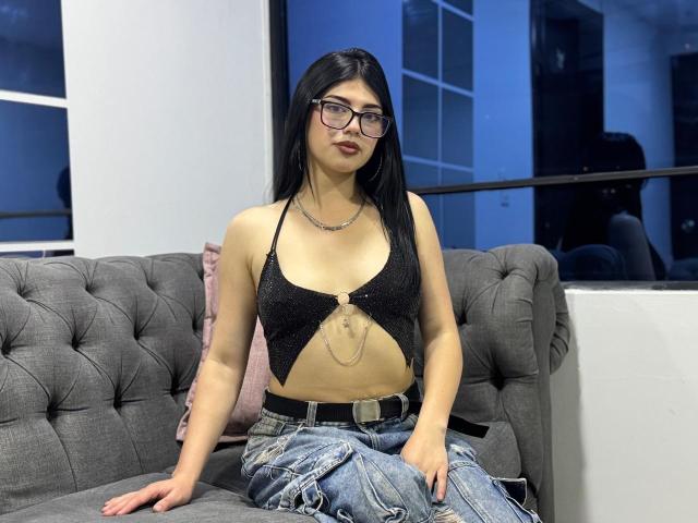 IsabellaaRossi with black hair, glasses, and dark lipstick, wearing a sparkly black top with cutout, chain detail, and blue jeans, sitting on a gray tufted sofa.