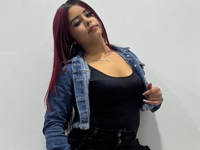 RominaRose with red hair, black tank top, and blue denim jacket, showing off her tits, wearing high-waisted black pants, against a white wall.