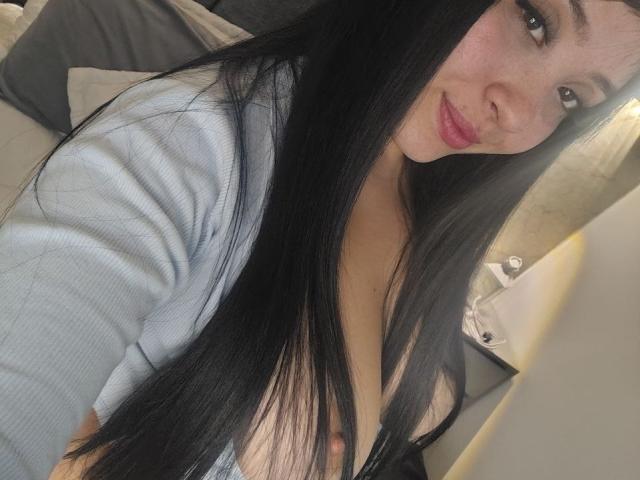 LunaBellaHotty with long black hair, light pink lipstick, and a grey shirt, showing her small tits, slightly visible under her shirt. She's smiling at the camera.