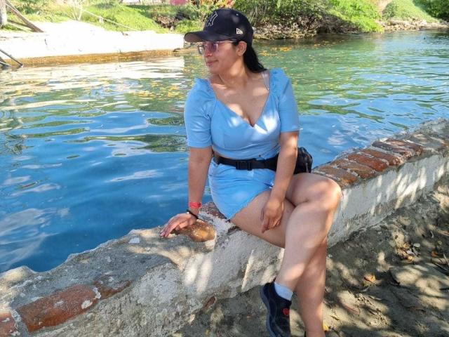 SofiiaThooner, wearing a blue dress and black cap, sits on a brick wall by a blue waterway, showing her tits in a low-cut top and black boots.