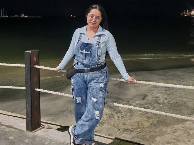 SofiiaThooner has titties, wearing ripped blue denim overalls, a light blue long-sleeve shirt, and glasses, standing by a rope near a dark, reflective water body.