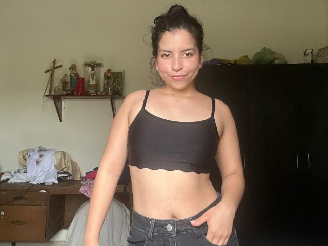 IanaPrince with dark hair in a bun, wearing a black sports bra showing her tits, and black jeans, standing in a room, smiling slightly, hand on her jeans.