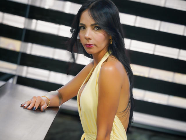 EvelinLuna with long black hair, yellow halter dress, backless, sitting at a table, looking over her shoulder, pink lipstick, gold earrings, bracelet, black nail polish.