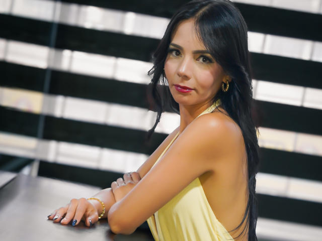 EvelinLuna with long black hair, wearing a yellow halter dress, sitting at a table, arms crossed, looking over shoulder, red lipstick, gold earrings, and a bracelet.