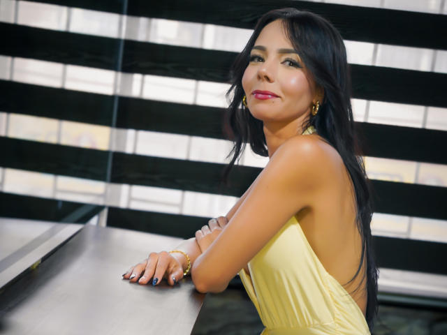 EvelinLuna with long black hair, yellow backless dress, pink lipstick, gold earrings, and bracelet, leaning on a table, smiling, showing her tanned ass.