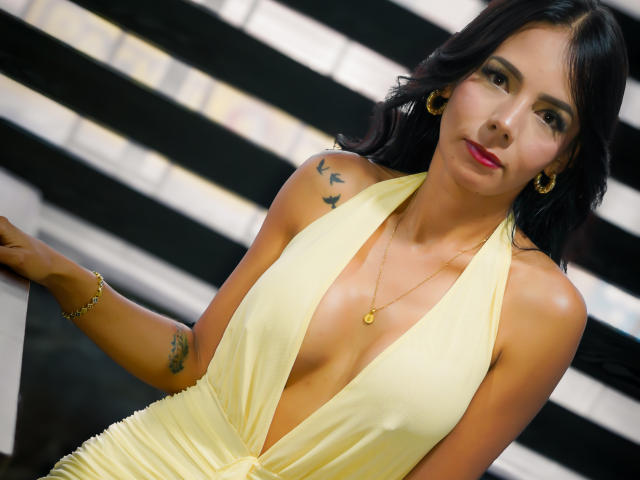 EvelinLuna with black hair, yellow halter dress, deep cleavage, gold earrings, necklace, tattoos on arm, leaning on black and white striped surface, looking at camera.