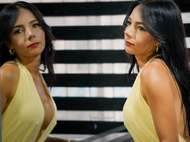EvelinLuna with long black hair, red lipstick, gold earrings, and a yellow halter dress, showing her large tits in a mirror reflection. Her ass is not visible.