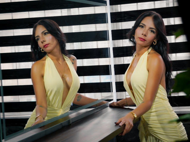 EvelinLuna with big tits in a yellow, deep-pleated dress, standing by a mirror, showing her cleavage, and looking at the camera, with black hair and red lipstick.