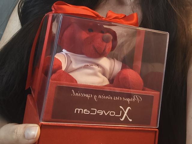 TaniaSandersX holding a red, stuffed teddy bear in a clear, red box with a red ribbon. Teddy wears a white shirt with "Jocelyn" embroidered. Her black hair visible.