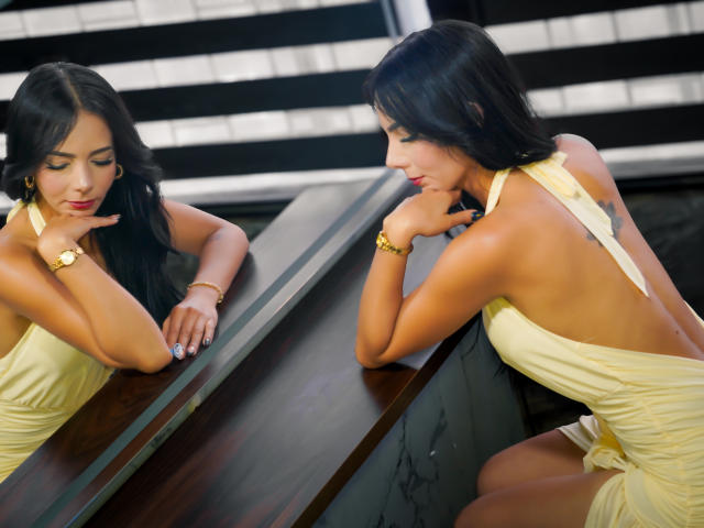 EvelinLuna with black hair, yellow halter dress, and gold bracelet, sitting by a mirror, looking at her reflection, touching her chin, showing her back and small tattoo.