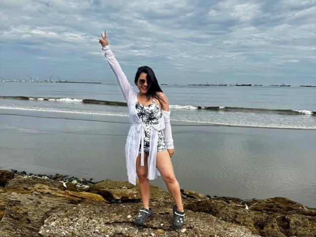 WendyStacye stands on rocky shoreline, arm raised in a peace sign. She wears a white floral dress with a loose, sheer cardigan. Black sneakers with white accents. Dark hair, sunglasses. Overcast sky, calm sea with small waves, distant ships. Her right leg is slightly forward, left leg back. Rocks and pebbles beneath her feet. She looks confident, smiling. The image has a casual, natural style. WendyStacye is centered, taking up most of the frame. The photo is candid and relaxed.