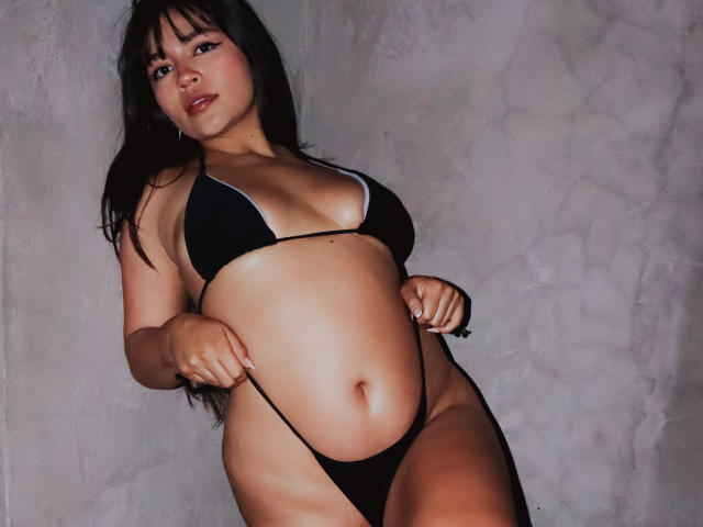 KimDevinne com biquíni preto, mostrando os seios e a vulva, puxando a tanga para baixo, em pé contra uma parede cinza texturizada, cabelos pretos longos, pele clara, olhando para a câmera..