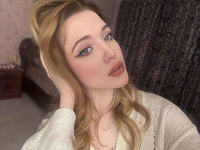 CarryParker, con cabello rubio claro, vestida con un suéter blanco de punto, tiene una mano en el cabello, ojos azules y un rubor rosado en las mejillas, mirando a la cámara..