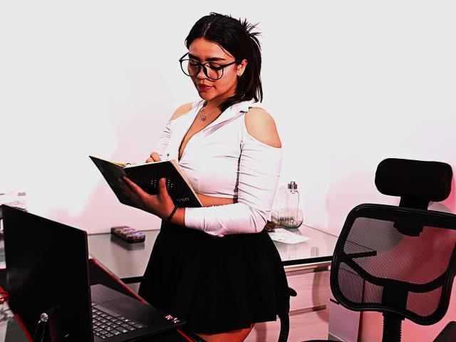 KimDevinne, com cabelo escuro, óculos, blusa branca com recortes, saia preta, segurando um tablet, em pé ao lado de uma mesa com um laptop, uma cadeira preta à direita, uma garrafa de água na mesa..