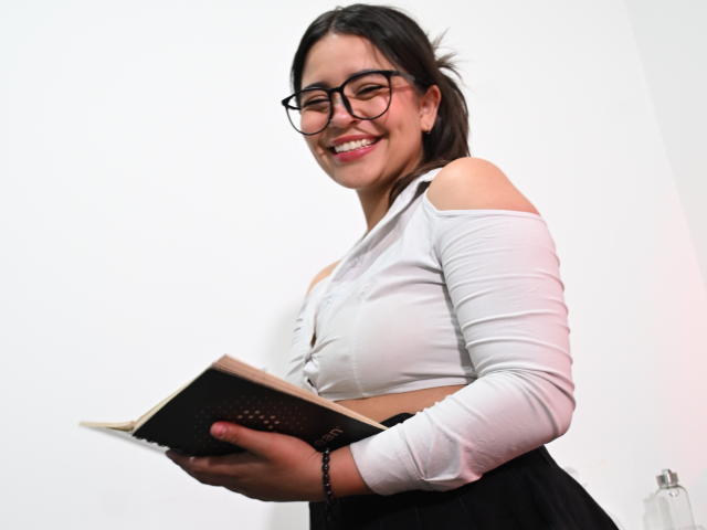 KimDevinne with glasses, smiling, wearing a white off-shoulder top, black skirt, holding a book, her small tits slightly visible through the top, dark hair in a ponytail.