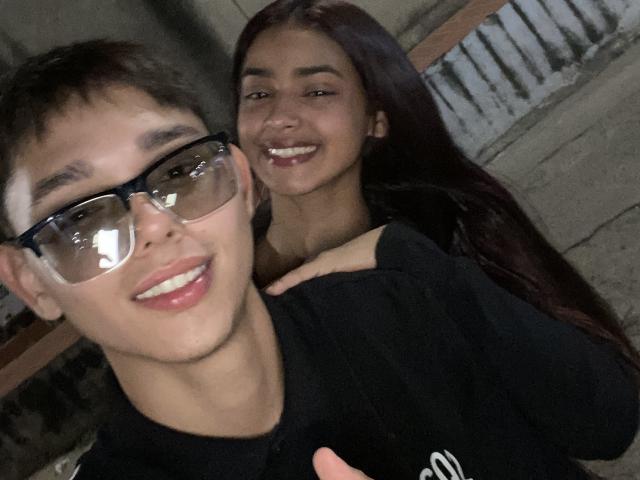 YouAndIsex with glasses and short brown hair, smiling at camera, has hand on YouAndIsex's black shirt. YouAndIsex with long dark hair, smiling, stands behind. Both look cute.