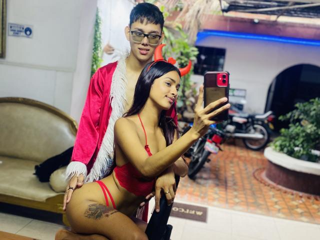 YouAndIsex in red lingerie, tits showing, tattoo on left hip, taking a selfie with YouAndIsex in pink jacket, glasses, and black hair standing behind her, holding her waist.
