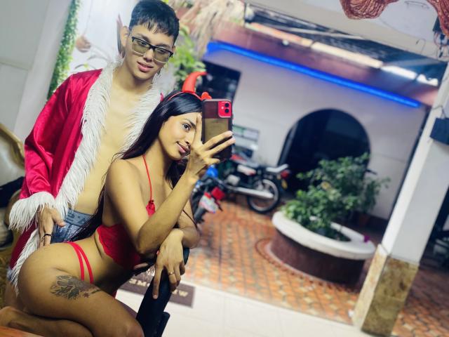 YouAndIsex with dark hair and red lingerie, showing her tits and tattoo, holding a phone, while a nerdy guy in a red robe and glasses stands behind her.
