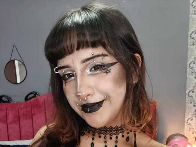 DanniLuke with dark bangs, goth makeup including black lipstick, pierced eyebrows, nose ring, and star-shaped eyeliner, wearing a black choker and lace necklace.