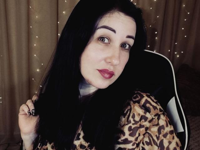 JillSilvies with long black hair, purple eyebrows, red lipstick, leopard print shirt, holding hair, seated in chair, fairy lights in background, looking at camera, medium shot.