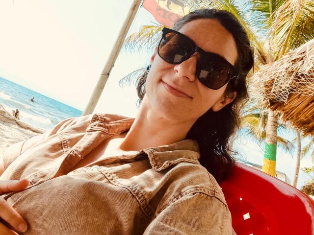 AndromedaJade has medium-length dark hair, wearing black sunglasses and a beige denim shirt. She's sitting on a red chair, leaning back with a slight smile. The image is brightly lit, with sunlight highlighting her face. She has a fair skin tone and is relaxed. A palm tree and beach are in the background. The composition focuses on her upper body and face. The texture of her shirt is slightly worn. The overall style is casual and summery.