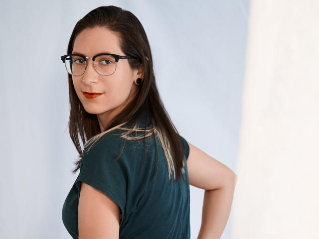 AndromedaJade with brown hair, glasses, and red lipstick, looking over her shoulder, wearing a green shirt, her hands on her hips, showing her tits slightly.