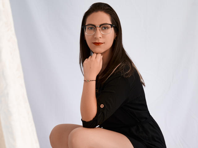 AndromedaJade with glasses, brown hair, black dress, red lipstick, squatting, right hand on chin, left arm on knee, wearing bracelet, white background, looking at camera.