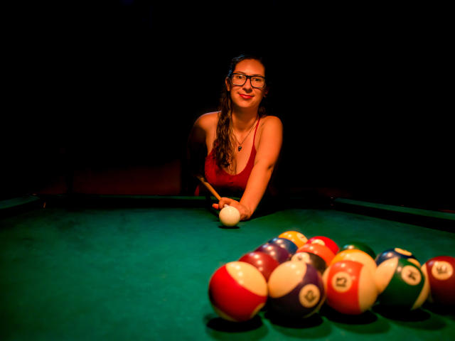 AndromedaJade with glasses, red tank top, and long brown hair, leaning on a pool table, holding a cue stick, smiling, her tits visible, in dark lighting.