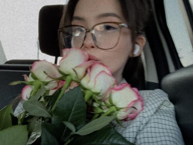 JeanieCat with glasses, holding pink and white roses to her mouth, wearing a checkered shirt, white earbuds, and brown hair, sitting in a car.