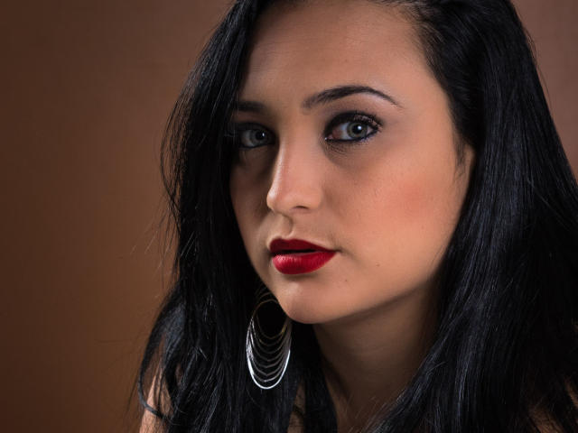 Close-up portrait of DanyQueenX. She has dark, wavy hair and a serious expression. Her bright blue eyes are slightly squinted, and she's wearing bold red lipstick. A large, silver, spiral earring hangs from her right ear. The brown background is plain and unobtrusive. The lighting is soft, highlighting her facial features and creating a slight shadow on her left cheek. The texture of her hair is smooth, contrasting with the matte finish of her skin.