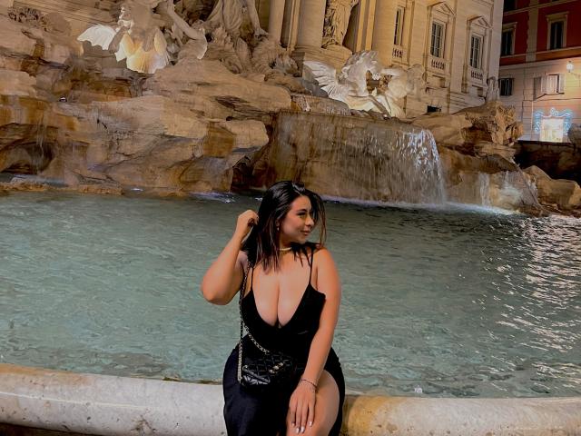 GabrielaFerrer with big tits in a black dress, sitting by a fountain, hand in hair, looking down, water in front, rocky background, night, Instagram watermark.