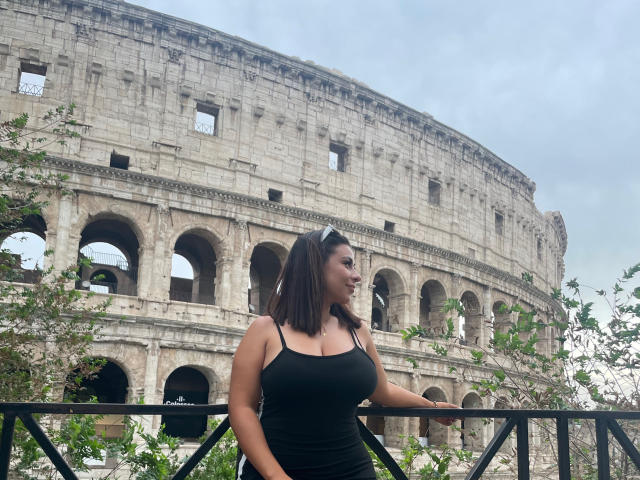 Gabriela Ferrer, met grote borsten, draagt een zwart tanktop en staat voor een vervallen Romeins Colosseum, met haar armen rustend op een zwarre reling, en groene bladeren op de voorgrond..