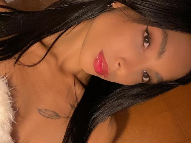 Close-up photo of AnnaDreams6. She has long, straight black hair, brown eyes, and full pink lips. Her skin is smooth and tan. She wears a fluffy white strap that reveals a small tattoo of a leaf on her right shoulder. The angle is slightly tilted, focusing on her face and upper chest. Her eyebrows are dark and well-defined. The lighting is warm, highlighting her features.