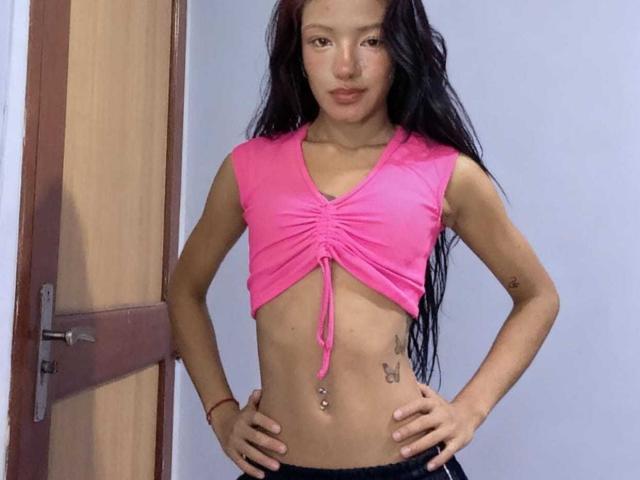 AnnaDreams69 has long black hair, brown eyes, pink glossy lips, and a small leaf tattoo on her chest, wearing a white furry strap. Her ass is not visible.
