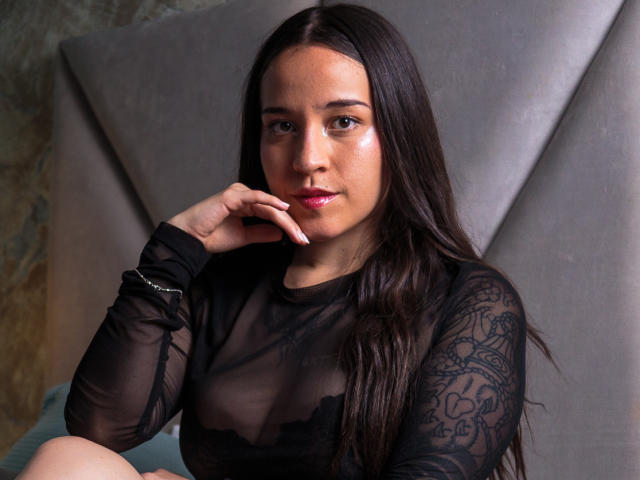 IrissMontiel, a young woman with long dark brown hair, sits against a gray, padded headboard. She wears a sheer black lace top, revealing her small breasts and a hint of her nipples. Her right hand rests on her chin, showing a silver bracelet. Her expression is neutral, with slightly parted pink lips. The lighting is soft, highlighting her smooth, slightly shiny skin. The image has a watermark in the bottom right corner.