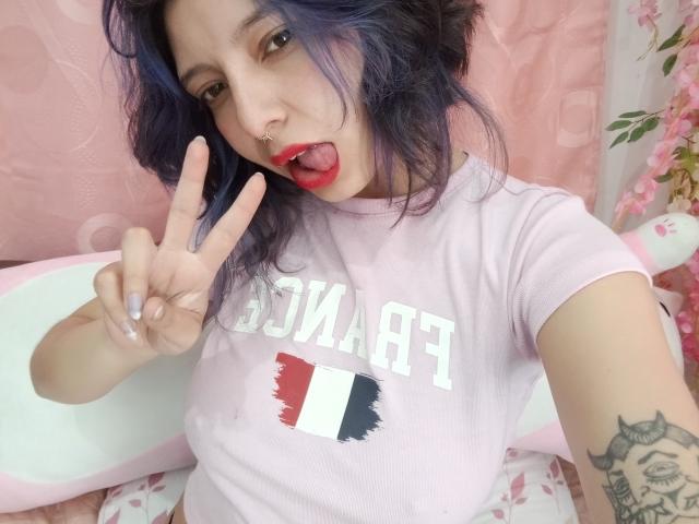 KiaraXMoon kneels on a gray bed, wearing a red sheer top and red thong, showing her ass, with long black hair, in a purple, padded wall room.