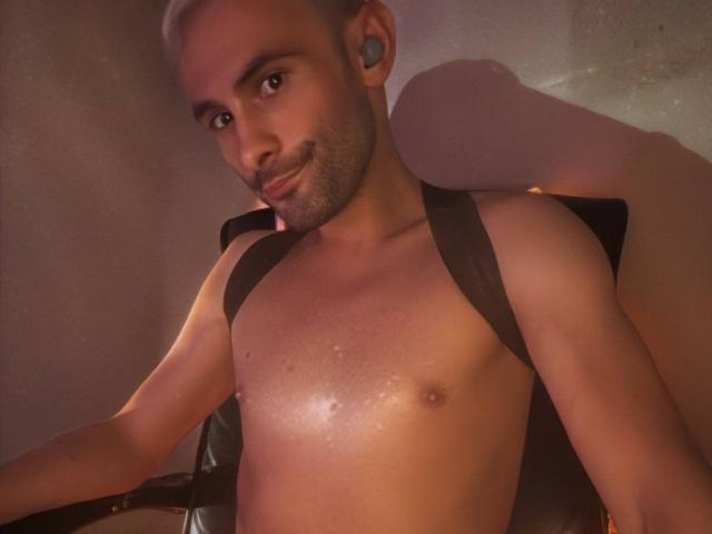 Un uomo senza camicia, con un sorriso carino, piccoli capezzoli, una leggera peluria sul petto, piercing neri alle orecchie, capelli biondi corti e una leggera barba, indossa bretelle nere, in un ambiente con scarsa illuminazione..