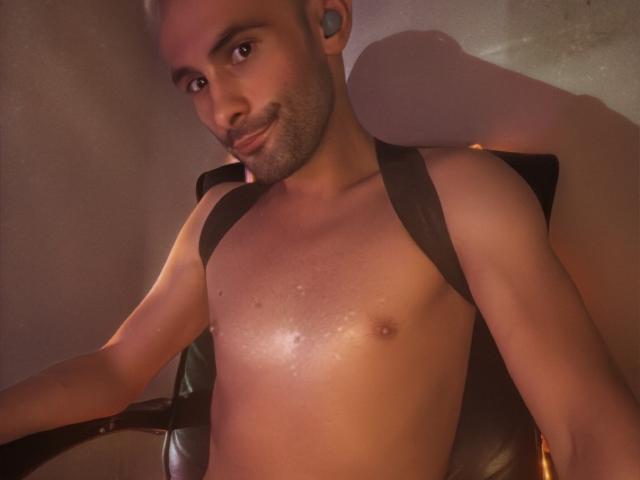 shirtless hTxmTx with a black harness, sitting in a chair, looking at the camera, slight smile, earbuds in, short-cropped hair, light skin, small nipples, slight chest sweat.