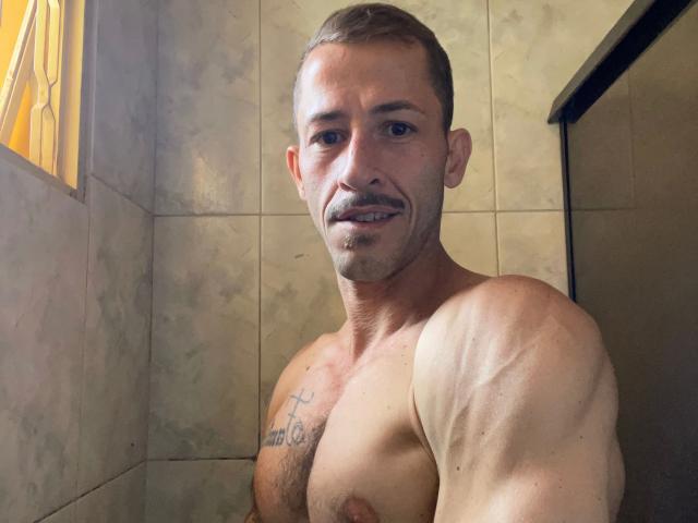 LucasJandre-attraktiv mit kurzen, braunen Haaren, grünen Brillen und einem weißen, strukturierten Hemd, trägt roten Lippenstift, hat einen tätowierten Arm, ein rotes Armband, sitzt im Auto, Sonnenlicht auf dem Gesicht, entspannt..