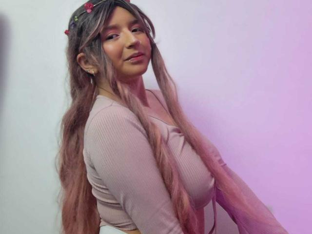 CandySwan with long pinkish-brown hair, wearing a pink ribbed top, black scrunchie on wrist, flower crown, resting chin on hand, soft pink and purple lighting.