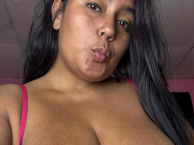 AnaisDelorme with dark hair, puffy brown lips, brown eyes, and tan skin, wearing a pink spaghetti strap top, showing her large, round boobs. Ceiling and pink wall in background.